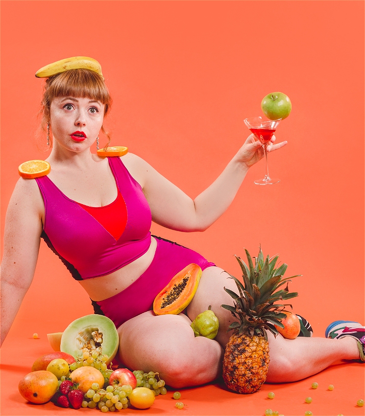 A woman wearing a two-piece pink and orange lycra outfit sits with fruit placed all around and on her, a banana is on her head. She has a surprised expression and holds a martini glass with an entire apple on the side.