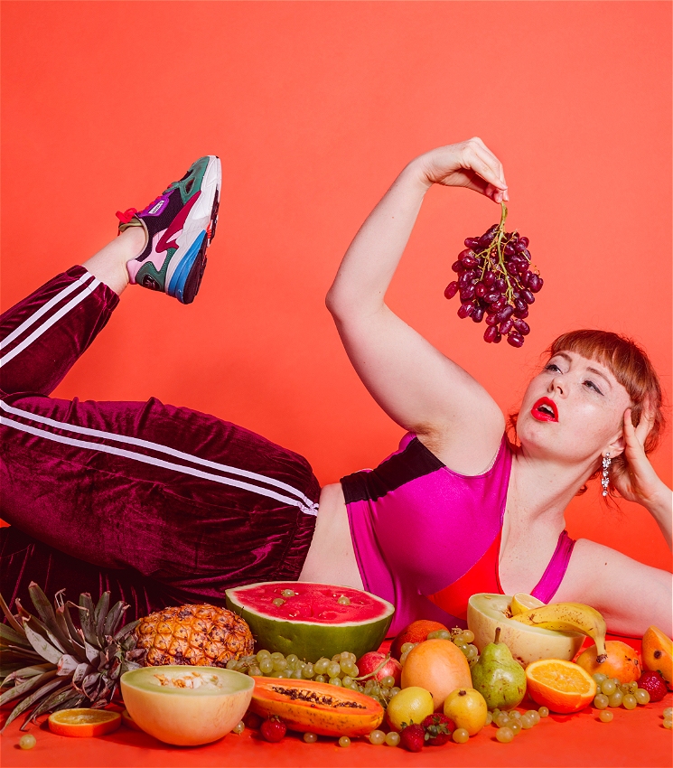 A woman lies on the ground amongst a lot of fruit, one leg is raised in the air in an athletic position, she rests her head on her hand. She is wearing a pink and orange lycra top and maroon velvet trackpants. She looks desiriously at the grapes she holds in her other hand.