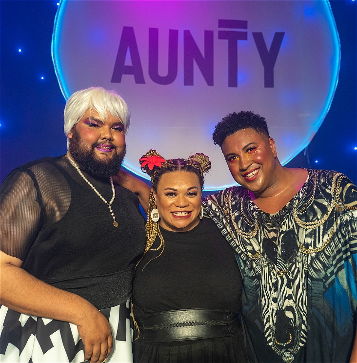 Three stylishly dressed people of colour embracing each other and smiling, standing in front of a blue background with star-like lights.